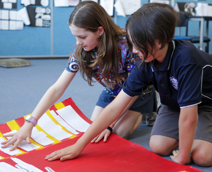 Taken in a bright classroom, two students are leaning over a red poster board, collaborating on a Science project.