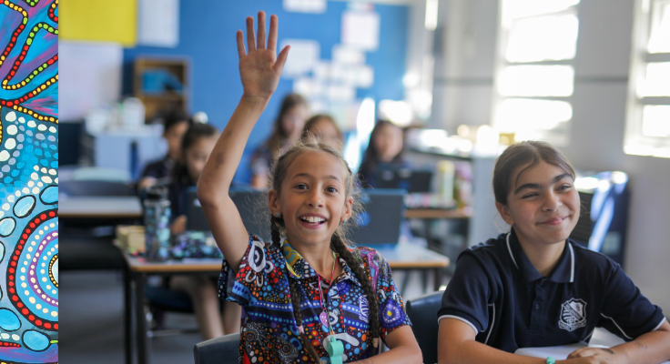 Two students sit in a bright, sunny classroom. The student on the left has her hand up, ready to answer a question. Both students are smiling.