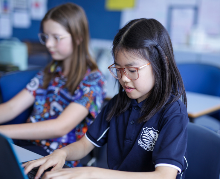 Two students are working on laptops in a bright classroom. One student wears the navy blue WPS shirt, the other wears the 'Dreaming' indigenous polo