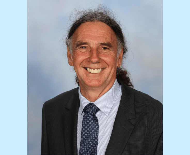 Portrait of Harold Cosier, principal of Wollongong Public School. He is smiling in front of a blue background.
