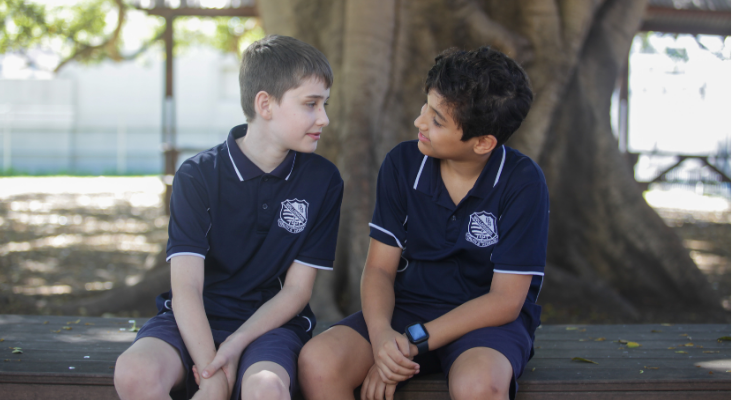 Two year 5 boys sit in the shade of the the WPS figtree. They are chatting to one another, smiling.