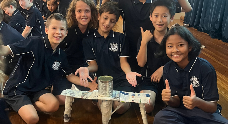 Five students, all wearing navy WPS polos, sit around a newspaper structure which is holding up a tin can. Some students are giving 'thumbs up'. They are smiling.