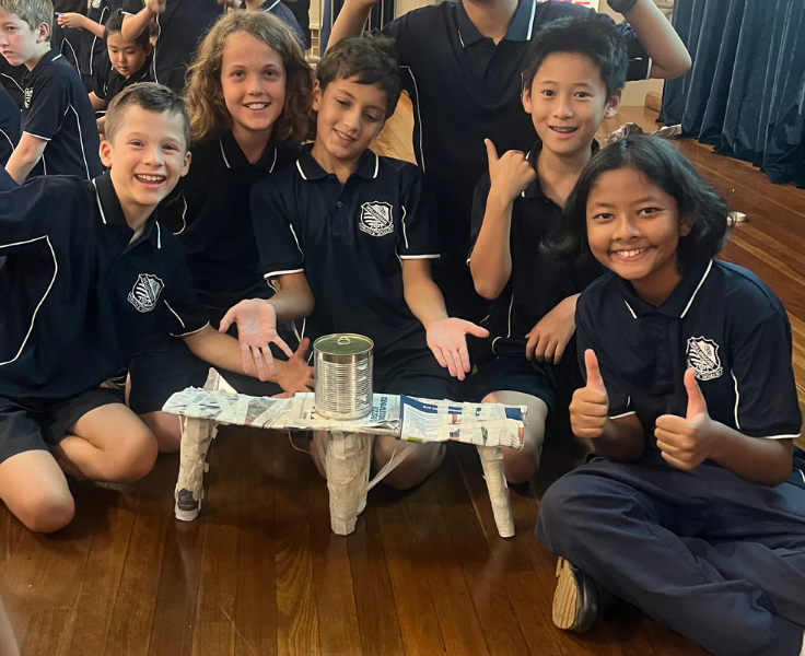 Five students, all wearing navy WPS polos, sit around a newspaper structure which is holding up a tin can during a STEM challenge. Some students are giving 'thumbs up'. They are smiling.