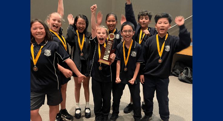 A group of eight students stand cheering and smiling, the one in the middle holds a large trophy. They each wear yellow and gold medals around their neck.